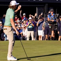 Professional golfer in a green shirt and cream pants holds up the golf ball to the spectators