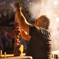 Chef skewers meat onto a long metal rod at tall as his head as smoke surround him at Tasting Australia