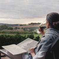 Woman sits outside sipping her red wine and reading a book with a view over the rolling vines in the Barossa