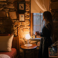 A woman stands by the bedroom window inside the cosy cottage as the lamplight glows warm inside. 