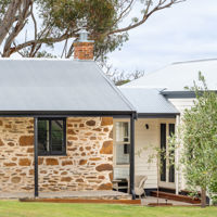 A stone cottage with a chimney called Merilli1885 is framed by a towering gumtree in the Fleurieu Peninsula. 