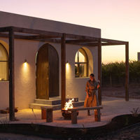 A girl walks to her seat outside with a wine in hand, where the fire is crackling and sun casts a purple glow