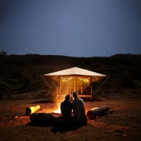Two friends sit closely on logs around an open fire with their retreat illuminated in front of them 
