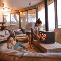 A couple sit in the lounge room, one sips her tea on the floor while her partner adds wood to the fireplace.