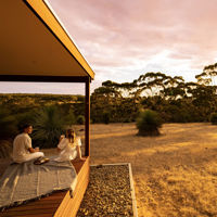 A couple sit on the decking drinking and eating while watching the sun set on the nature around them