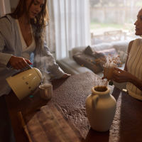 Two women chat around a kitchen bench while pouring cups of hot tea as light spills through the window 