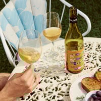 A bottle of their alphabet wine sits on a white table with two full glasses and a side of bread and olives