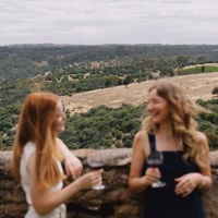 Two women drinking red wine against the backdrop of a sweeping view of rolling paddocks behind them
