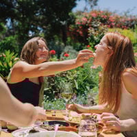 Friends sitting around a table outside filled with local produce as one girl playfully feeds her friend