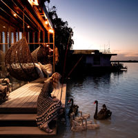A woman sits on the deck of The Cube, greeted by a black swan with water surrounding the accommodation