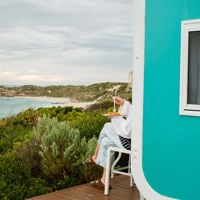 A woman eats fish and chips on the deck of her Hollocks Ocean Pod looking out toward the beach