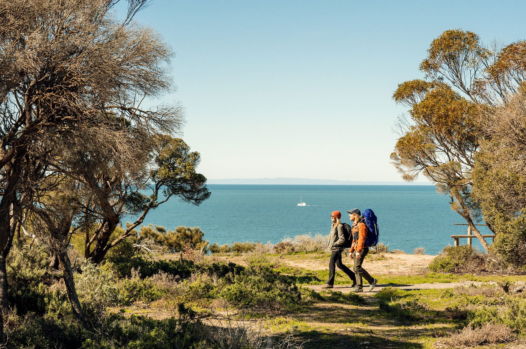 Two hikers with backpacks walking along a grassy trail surrounded by coastal trees, overlooking calm blue waters with a sailboat in the distance.